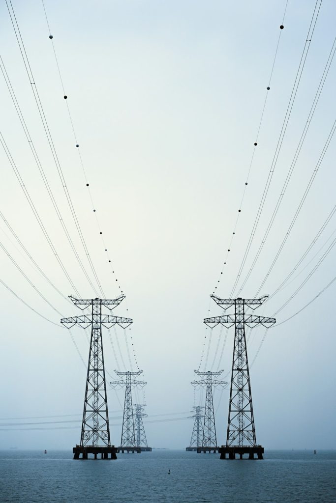 Two lattice electricity pylons carry a cross-sea power line over the calm waters of Shenzhen Bay, Guangdong, China. Soft morning haze flattens the colour palette, turning the steel towers and sweeping cables into a minimalist industrial seascape with strong leading lines.
